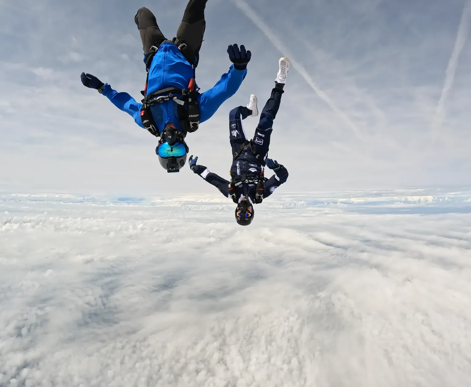 Deux parachutistes en combinaison bleue, tête en bas, équipés de systèmes d’oxygène, chutent librement au-dessus des nuages à 7700 mètres d’altitude.