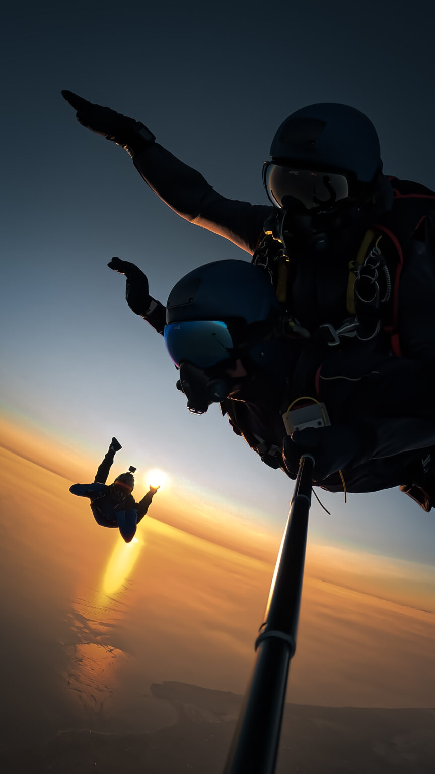 Deux parachutistes en tandem en chute libre au crépuscule, capturés par une perche selfie. Un troisième parachutiste apparaît en arrière-plan, semblant