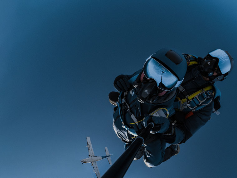 Tandem en chute libre à très haute altitude lors d'un saut HALO à 7 700 mètres — ciel bleu nuit d'altitude, moniteur et tandemiste équipés de combinaisons intégrales et masques à oxygène, avion de largage visible en contrebas, Vendée Évasion, Sables-d'Olonne