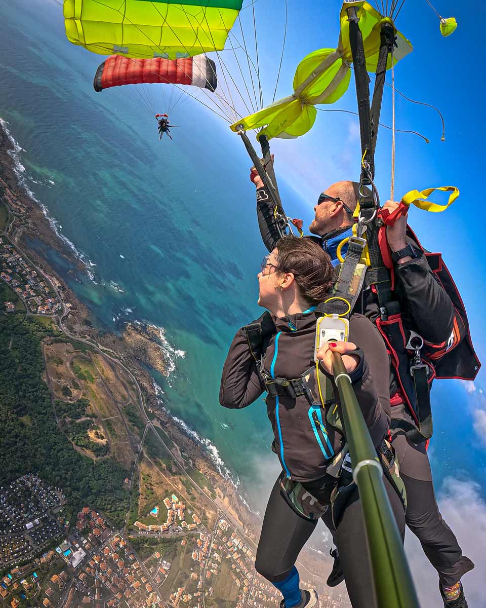 Tandemiste et moniteur Vendée Évasion en descente sous voile jaune-verte au-dessus des falaises et du littoral vendéen — un second parachute rouge visible en arrière-plan, vue sur l'Atlantique depuis 1 500 mètres, Sables-d'Olonne