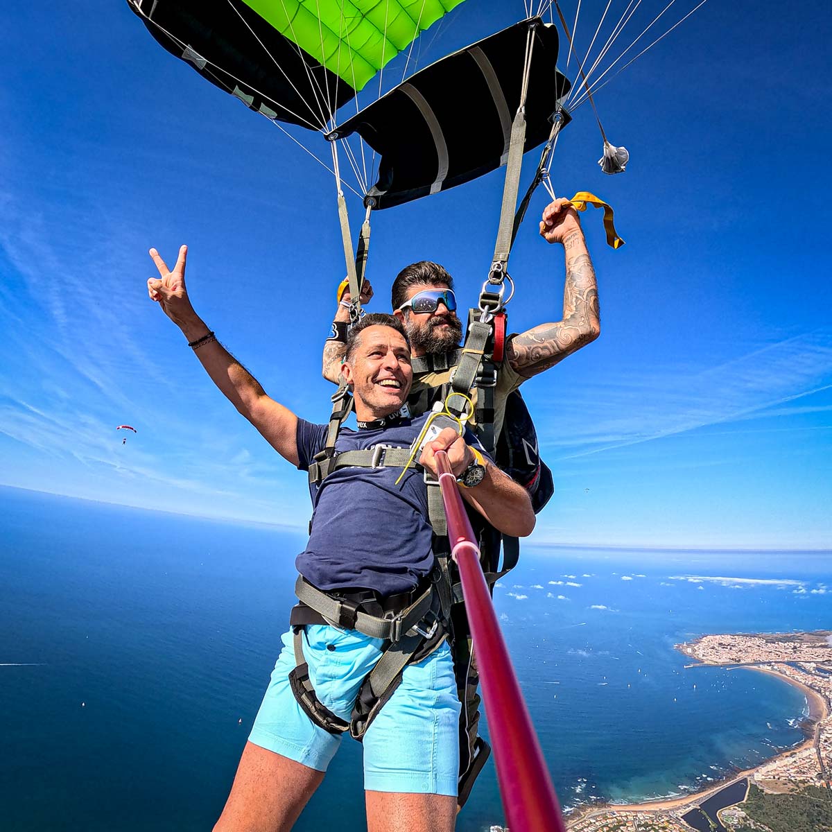 Saut en parachute tandem sous voile au‑dessus de la mer, passager et moniteur souriants avec la côte des Sables‑d’Olonne visible en arrière‑plan.
