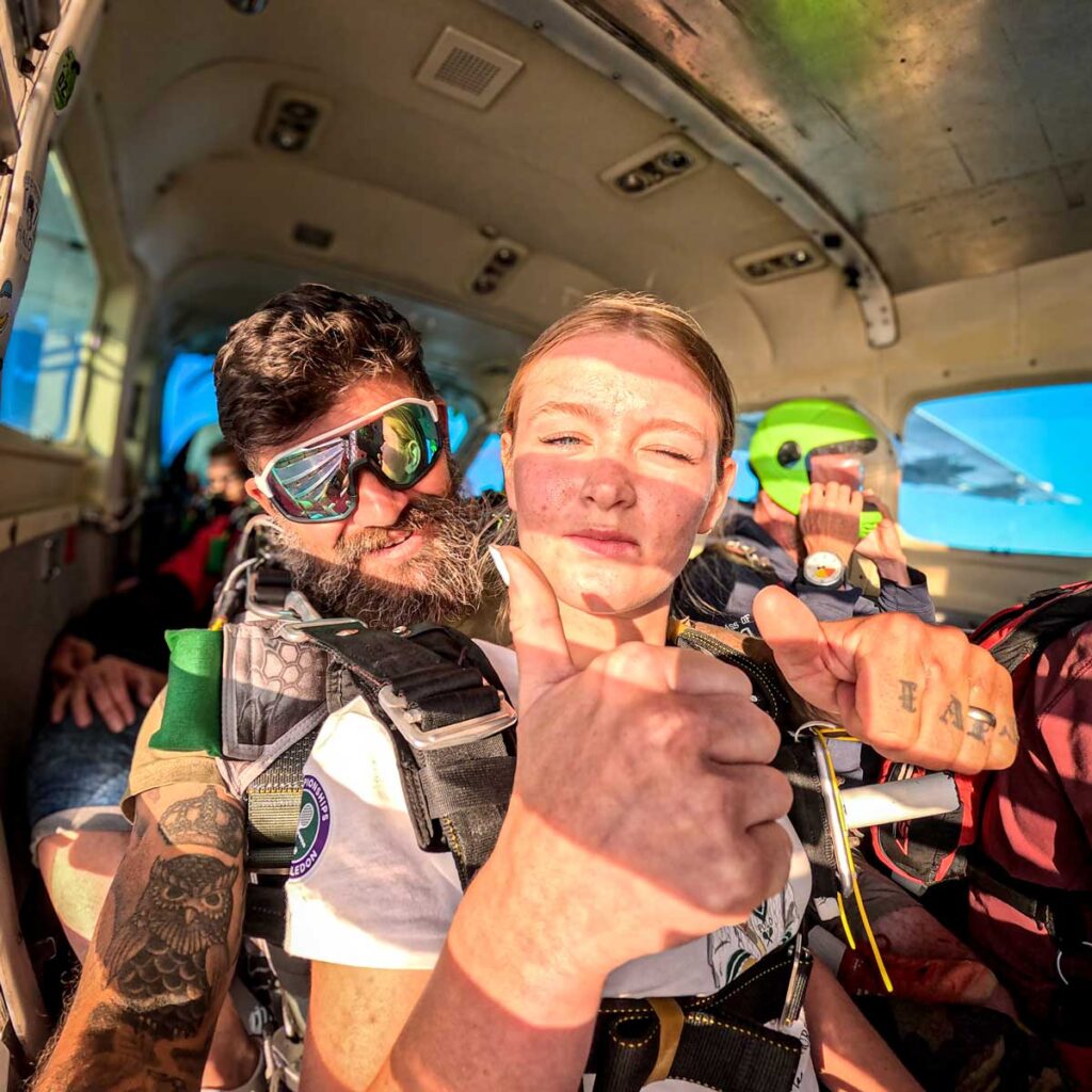Passager et moniteur de parachutisme assis dans un avion avant un saut tandem, souriants et faisant un signe positif avec le pouce.