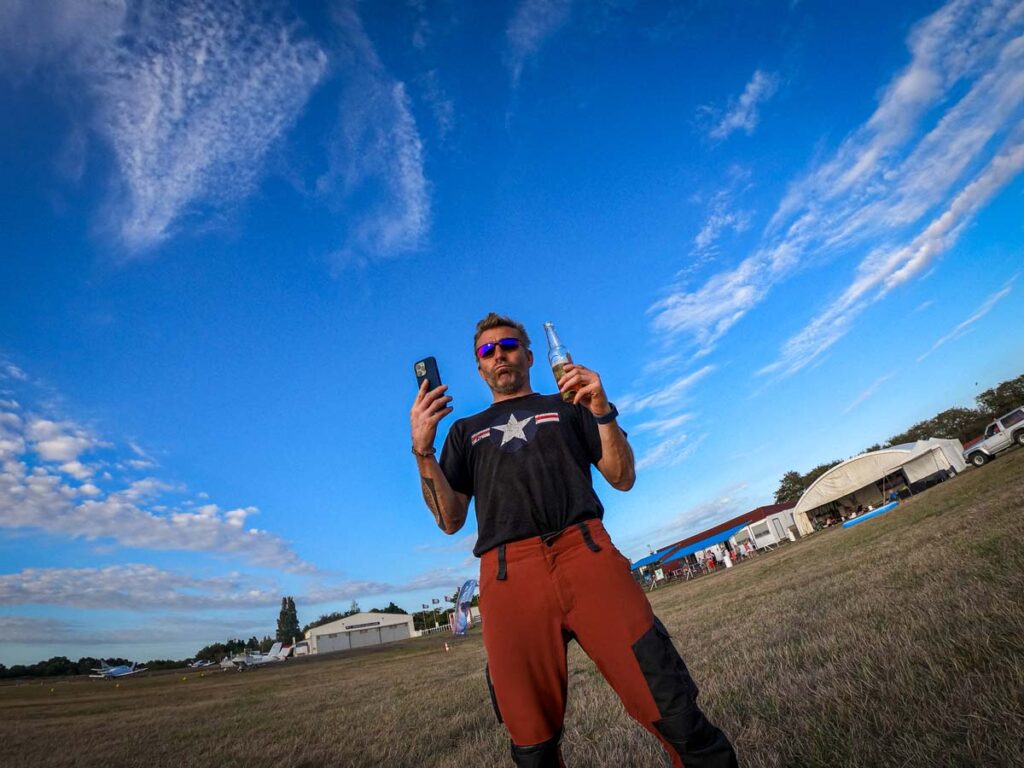Ludovic debout sur un aérodrome, tenant un téléphone et une boisson, sous un ciel bleu avec des nuages en arrière‑plan.