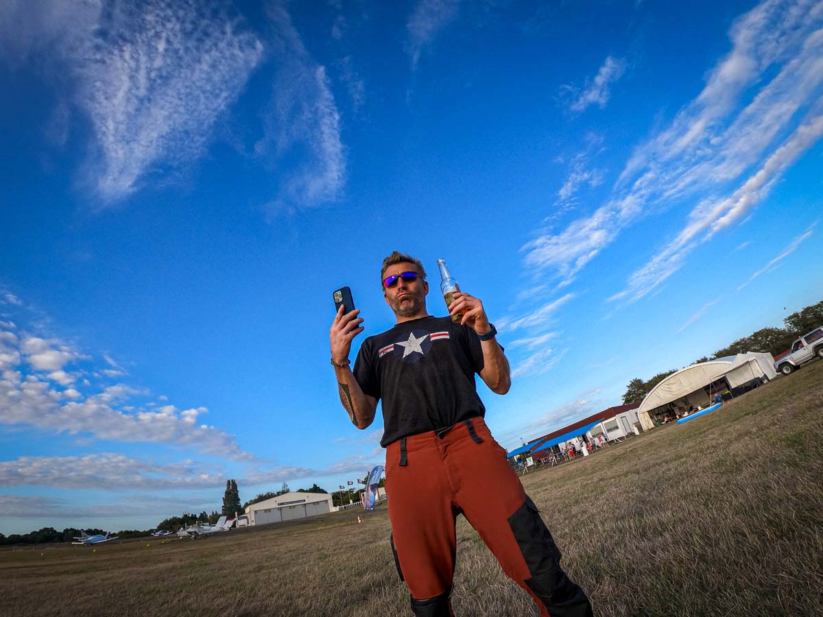 Ludovic debout sur un aérodrome, tenant un téléphone et une boisson, sous un ciel bleu avec des nuages en arrière‑plan.