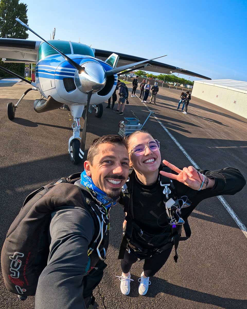 Moniteur tandem et tandemiste souriants en selfie sur le tarmac de l'aérodrome des Sables-d'Olonne, harnais tandem enfilés, devant le Cessna 208 Caravan de Vendée Évasion — avant l'embarquement pour le saut en parachute